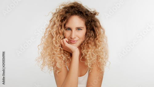 head and shoulders plan of blonde wooman with big curly hair in studio on white watching directly to the camera has a sort of flirt look