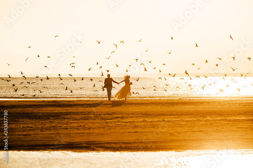 Silhouettes of the bride and groom, holding hands, run on the beach at sunset on the background of a flying flock of birds.