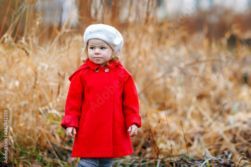Outdoor portrait of little cute toddler girl in red coat and white fashion hat barret. Healthy happy baby child walking in the park on cold day. Fashion stylish clothes for kids. © Irina Schmidt