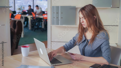 Red-haired adult woman at the office odering on laptop some goods. She holding orange debit card and shopping at the internet shop photo