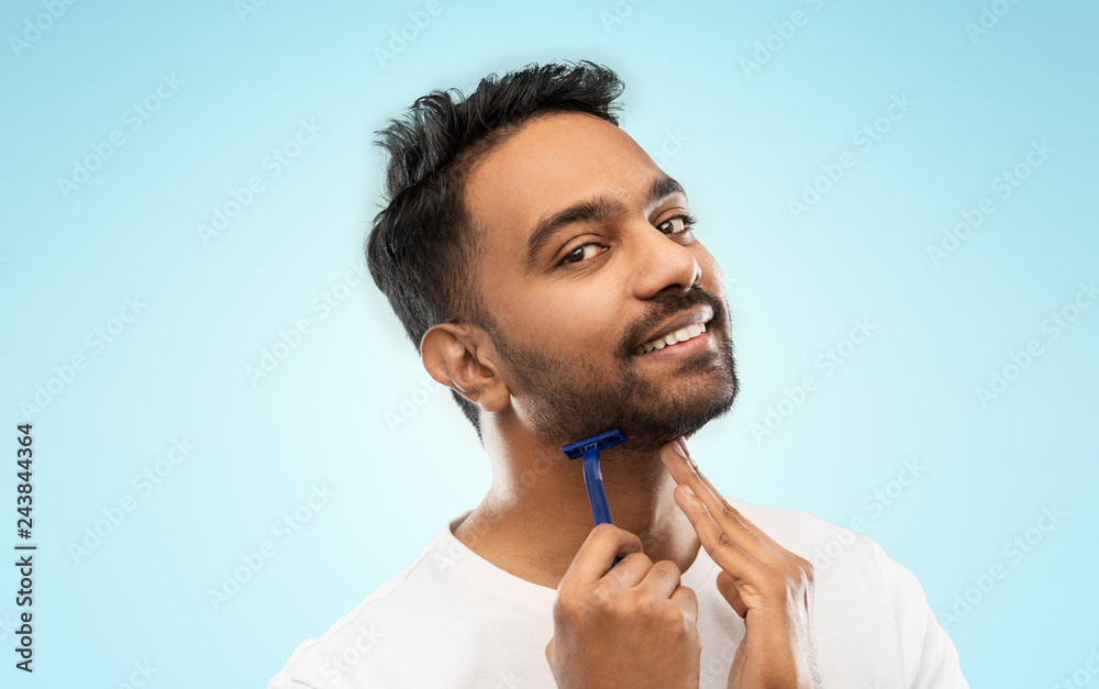 grooming and people concept - smiling indian man shaving beard with manual razor blade over blue background grooming and people concept - smiling indian man shaving beard with manual razor blade over blue background