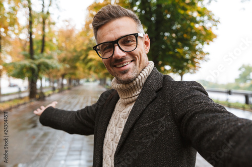 Photo of joyful man 30s wearing warm clothes smiling, while walking outdoor through autumn park