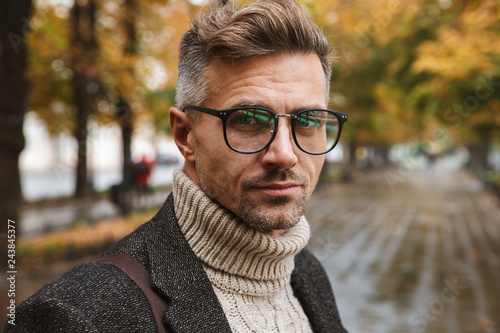 Photo of adult man 30s wearing eyeglasses looking at camera, while walking outdoor through autumn park
