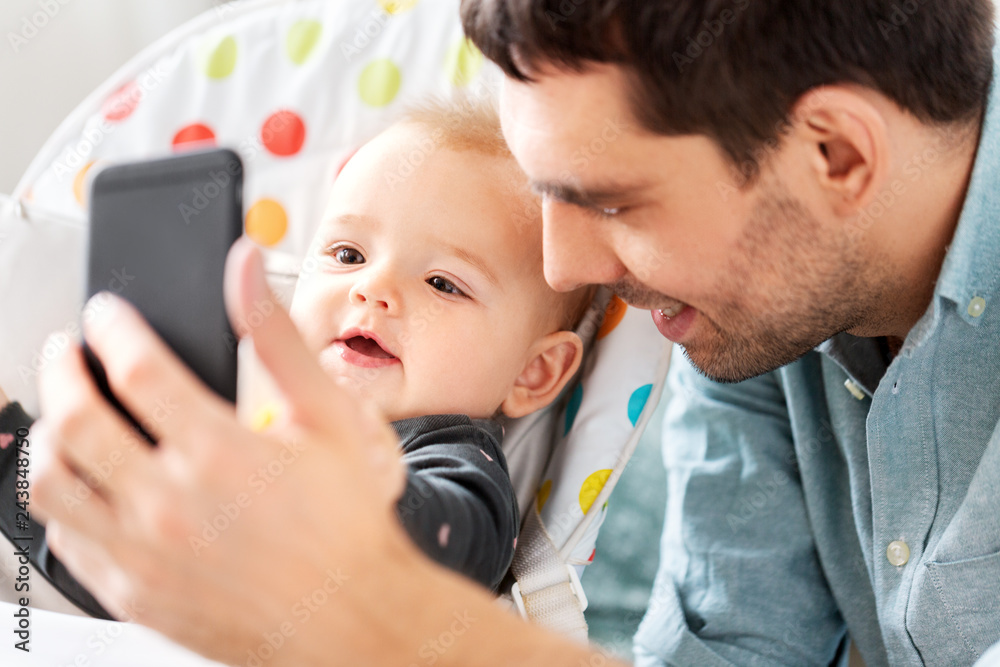 family, fatherhood and people concept - father with little baby daughter in highchair taking selfie by smartphone at home