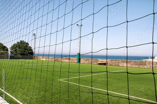 Look through net to empty football field in soccer stadium. Soccer field with green grass in football sport stadium behind net curtain. Sport equipment  football goal  white line  soccer field corner.