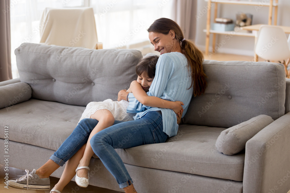 Happy mother hugging cute little girl sitting together on sofa in living room, smiling mom embracing kid, mum and daughter cuddle having fun at home, sincere warm relationships of mommy child concept