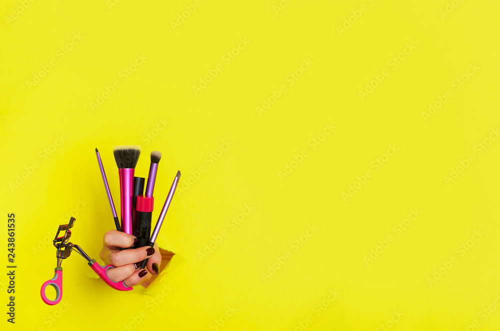 Woman hand with professional cosmetic tools for make up: brushes, mascara, lipstick, eyelash curler on yellow background. Beauty concept. Banner for cosmetics sale. Copy space