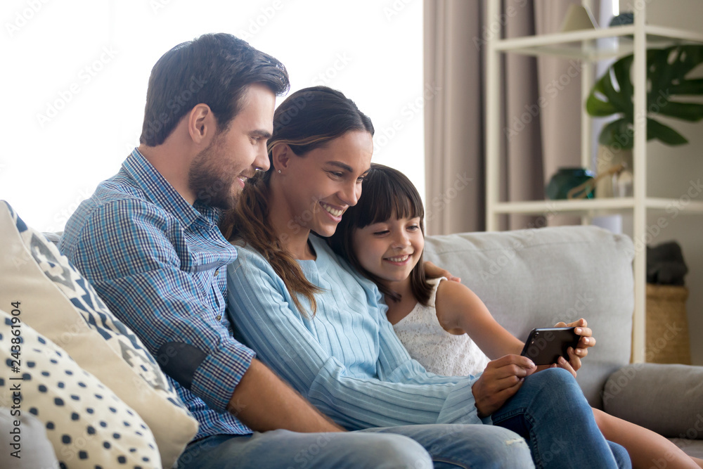 Beautiful family of mom dad and little kid child daughter sitting together on sofa smiling looking at smartphone screen taking selfie, making video call or recording vlog with cellphone at home Beautiful family of mom dad and little kid child daughter sitting together on sofa smiling looking at smartphone screen taking selfie, making video call or recording vlog with cellphone at home
