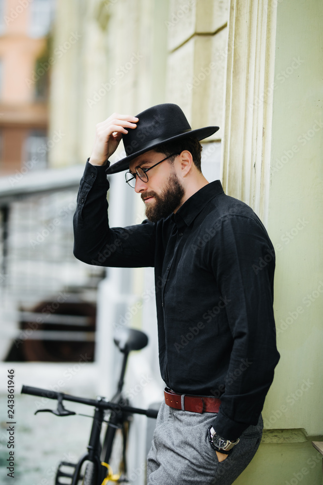 Street style. Pensive young bearded hipster man in hat with bicycle and looking away while standing on city street Street style. Pensive young bearded hipster man in hat with bicycle and looking away while standing on city street
