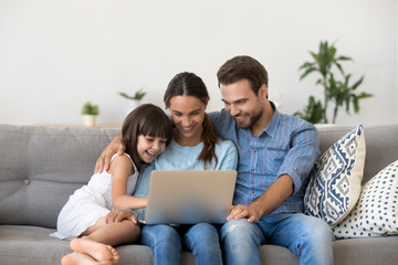 Happy family with kid girl having fun using laptop together sitting on sofa, parents and child daughter laughing relaxing at home with computer watching funny internet video, making online call