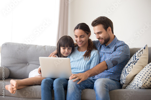 Happy family with kid daughter enjoy using laptop apps, making online call together, smiling parents and child girl relaxing having fun watching video on computer, doing internet shopping at home