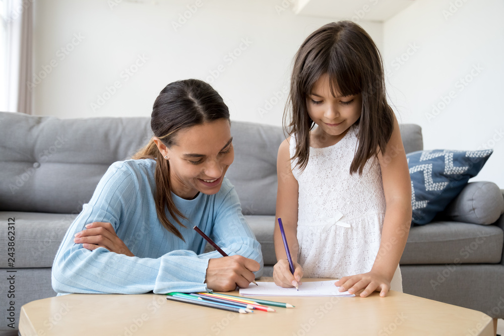 Cute child girl enjoys drawing with colored pencil on paper, mom or baby sitter helps kid teaching little daughter learning to draw at table play having fun with mother together at home or in daycare Cute child girl enjoys drawing with colored pencil on paper, mom or baby sitter helps kid teaching little daughter learning to draw at table play having fun with mother together at home or in daycare