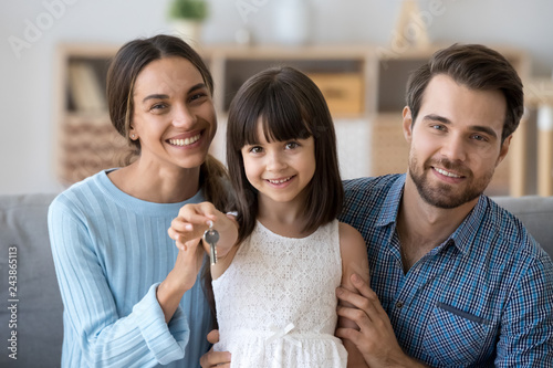 Cute kid daughter holding keys from new house looking at camera with mom and dad, child girl and parents home owners enjoying buying flat, happy family mortgage, real estate ownership, portrait
