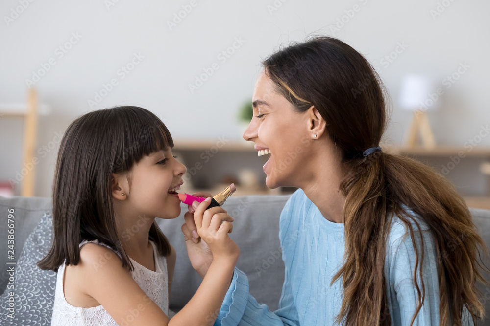 Happy mom and kid preschool daughter doing makeup together put lipstick on, funny little girl applying make-up on mothers lips, child having fun playing with mommy sister sitting on couch at home