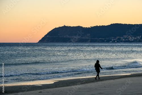 Woman with hooded parka walking on the sea shore of a sandy beach with a cape on the horizon at sunset, Alassio, Capo Mele, Liguria, Italy