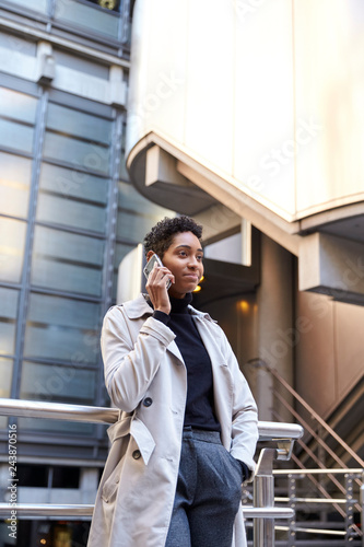 Fashionable young black businesswoman standing against handrail in the city talking on smartphone, vertical