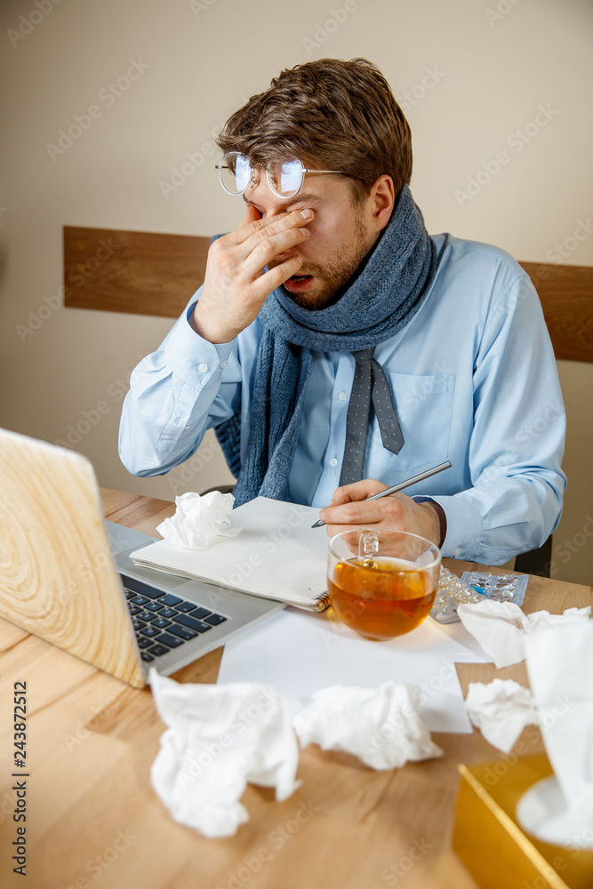 Feeling sick and tired. Frustrated sad unhappy sick young man massaging his eyes while sitting at his working place in office. The seasonal flu, pandemic influenza, disease prevention concept Feeling sick and tired. Frustrated sad unhappy sick young man massaging his eyes while sitting at his working place in office. The seasonal flu, pandemic influenza, disease prevention concept