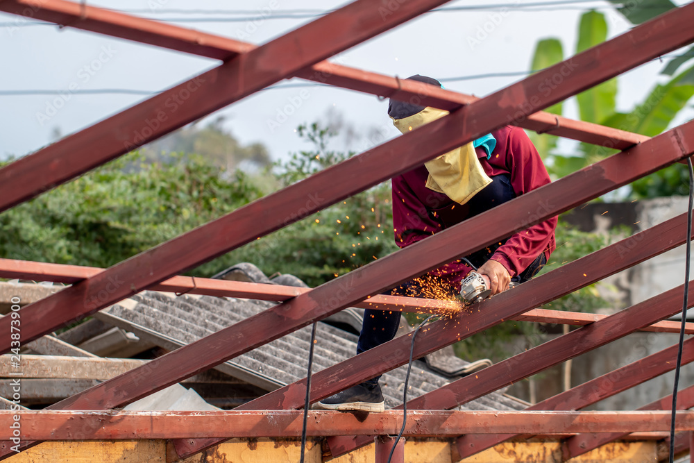 The man wearing brown shirt is cutting or sawing the metal on the roof construction by the metal saw on the top of the house in the sunny day. The man wearing brown shirt is cutting or sawing the metal on the roof construction by the metal saw on the top of the house in the sunny day.