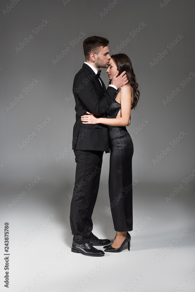 young tender man kissing forehead of beautiful girlfriend on grey