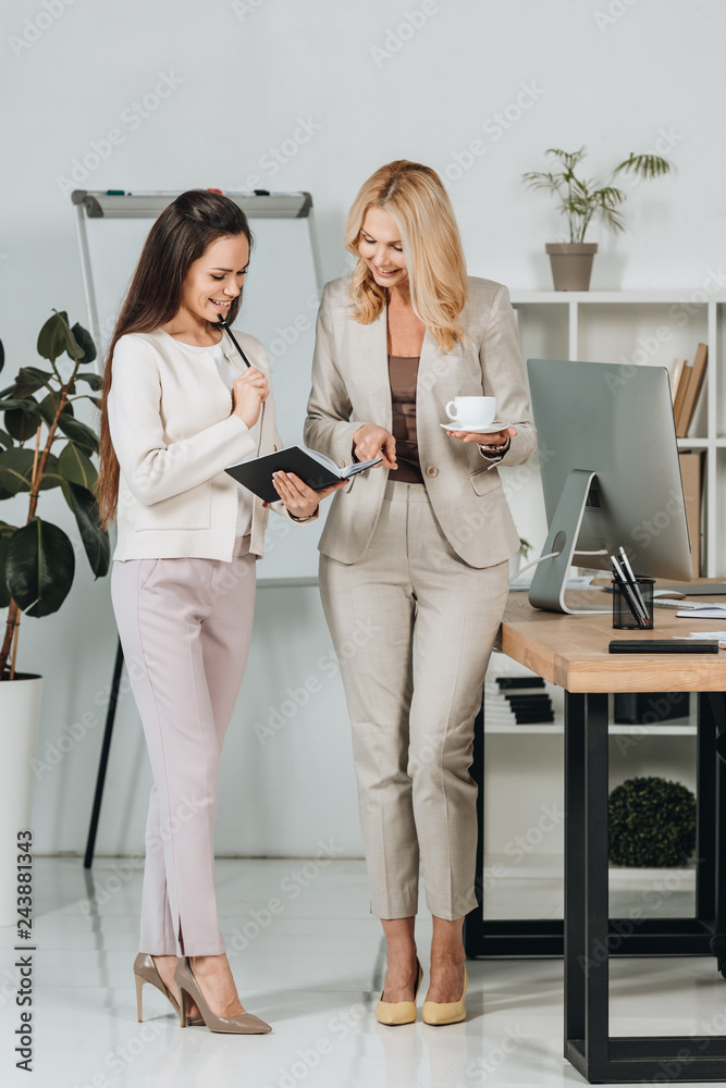 beautiful happy businesswomen with cup of coffee and notebook standing together and discussing work in office beautiful happy businesswomen with cup of coffee and notebook standing together and discussing work in office