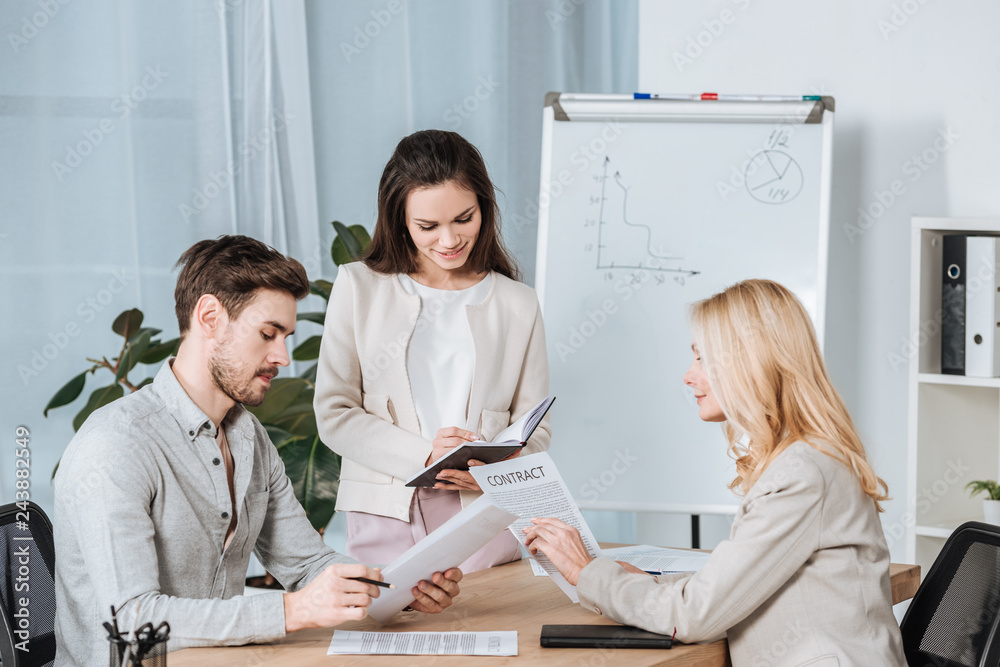 Fototapeta premium smiling young businesswoman writing in notebook and colleagues working with papers in office