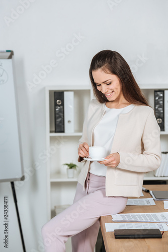smiling young businesswoman sitting on desk and looking at cup of coffee in office