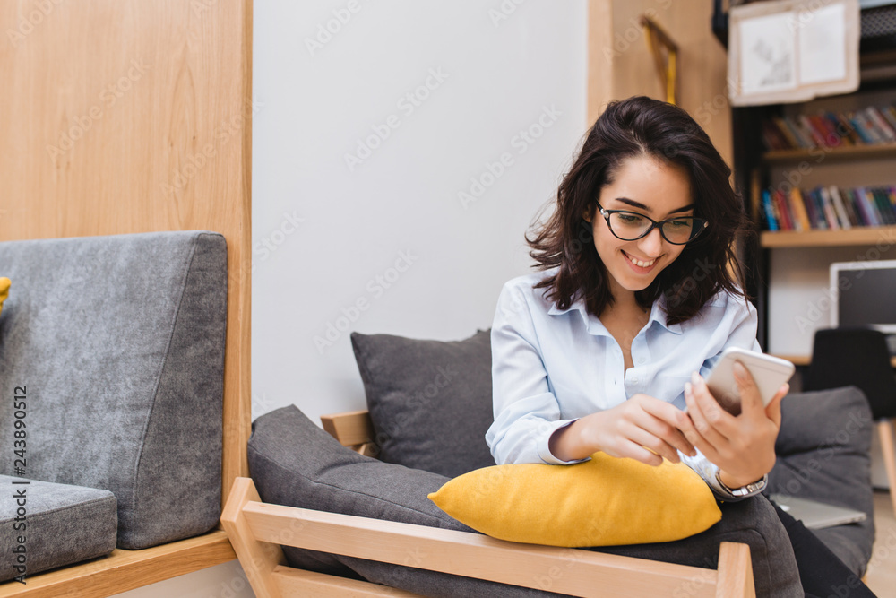 Portrait young joyful brunette woman in black glasses chilling on couch in modern apartment. Using phone, texting, smiling, cheerful mood. Place for text Portrait young joyful brunette woman in black glasses chilling on couch in modern apartment. Using phone, texting, smiling, cheerful mood. Place for text