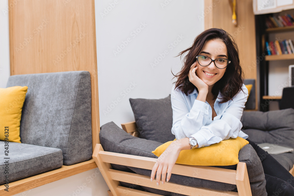 Portrait young brunette woman in black glasses chilling on couch in modern apartment. Comfortable, cheerful mood, smiling to camera. Place for text Portrait young brunette woman in black glasses chilling on couch in modern apartment. Comfortable, cheerful mood, smiling to camera. Place for text