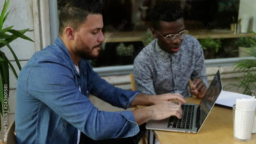 Multiethnic freelancers using laptop and discussing work. Focused young men working with laptop and talking while sitting together in outdoor cafe. Freelance concept Multiethnic freelancers using laptop and discussing work. Focused young men working with laptop and talking while sitting together in outdoor cafe. Freelance concept
