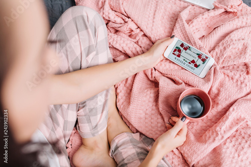 Home comfort on bed in modern apartment of pretty woman watching film on phone. View from above woman with cup of coffee enjoying domestic harmony, weekends, good morning on pink blanket