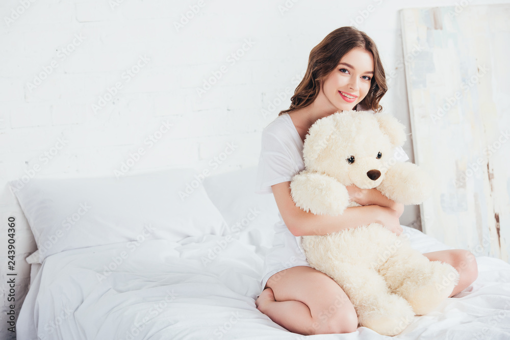 beautiful woman hugging teddy bear and sitting on bed with white bedding