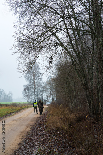 cloudy, foggy autumn day during hunting; rural road at the edge of the forest with two hunters who go away