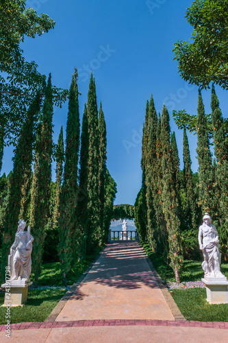 Beautiful white Italian sculptures in a hall surrounded by trees heading to the entry of a garden on blue sky.