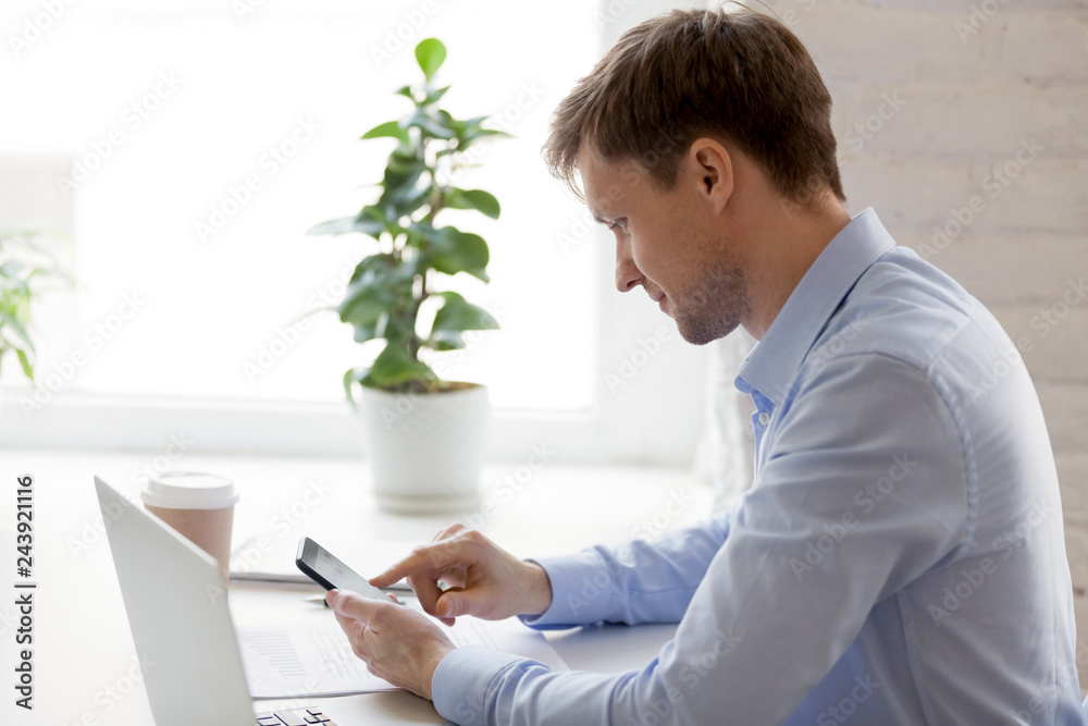 Confident focused businessman using phone and laptop at workplace, handsome man browsing mobile device apps, checking social networks, email, reading news, looking at smartphone screen in office Confident focused businessman using phone and laptop at workplace, handsome man browsing mobile device apps, checking social networks, email, reading news, looking at smartphone screen in office