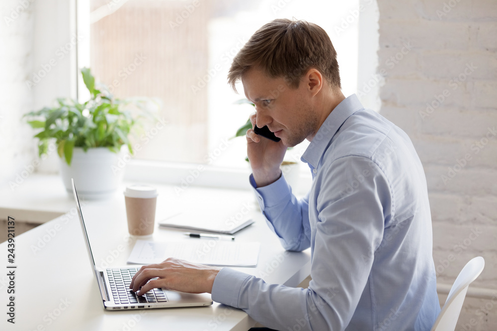 Focused businessman talking on phone and using laptop, serious man looking at computer screen, searching information, consulting client by smartphone, making call on workplace, working at project