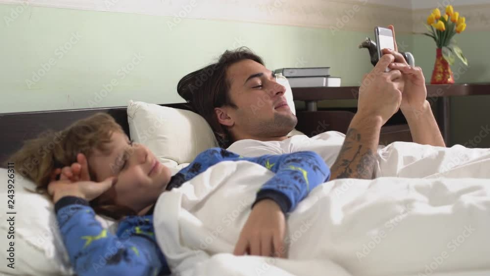 Two brothers lie down in bed covered with blanket, older guy chatting at mobile phone and laugh. Brothers relationship. Two brothers lie down in bed covered with blanket, older guy chatting at mobile phone and laugh. Brothers relationship.
