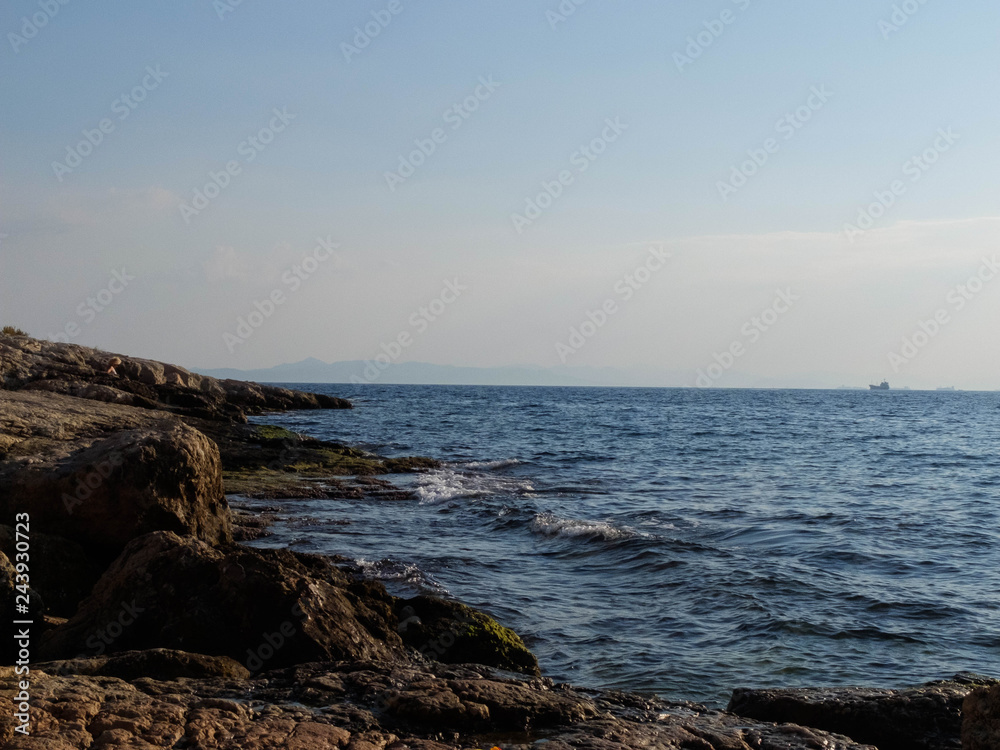 Rocky seashore, splashing waves and ships on the horizon. Piraeus, Greece