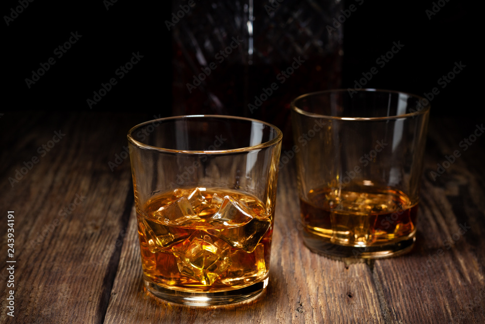 Two glass of whiskey with ice and crystal decanter on wooden table.