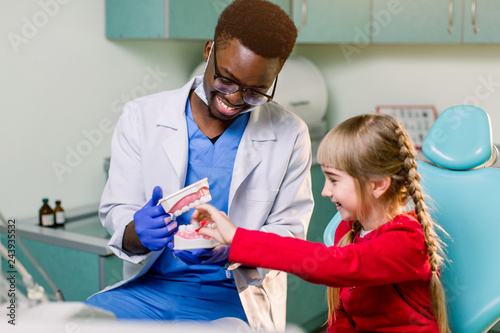 African dentist shows how to brush teeth to a small Caucasian girl a patient, using a artificial jaws. Dentistry, teeth hygiene concept