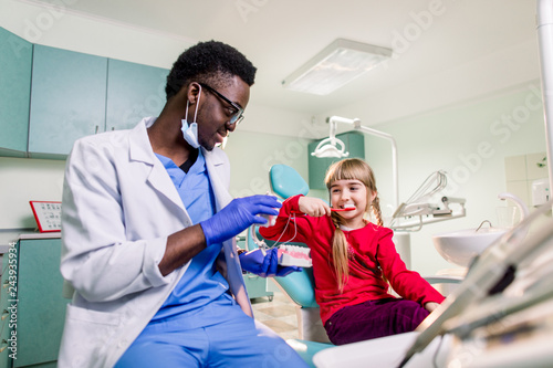 Little Caucasian girl is sitting in dental chair and smiling to African American dentist with artificial jaw in his hands