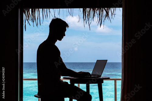 silhouette of a young man working with a computer on a table. Clear blue tropical water as background