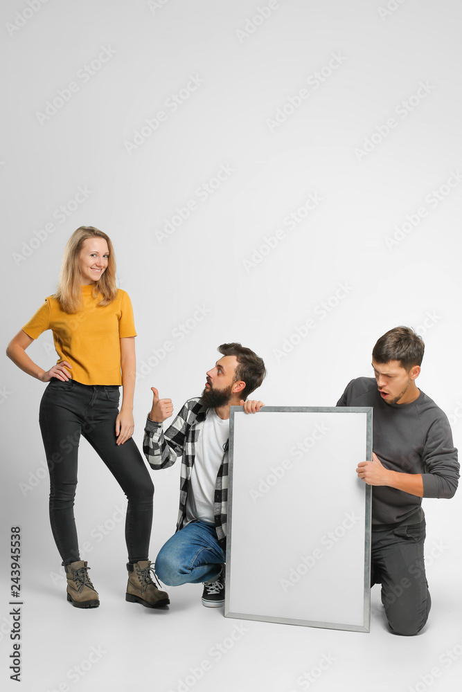 three funny laughing young people wearing casual fashionable clothes, on hunkers, posing, holding mockup big white board isolated in studio, white background