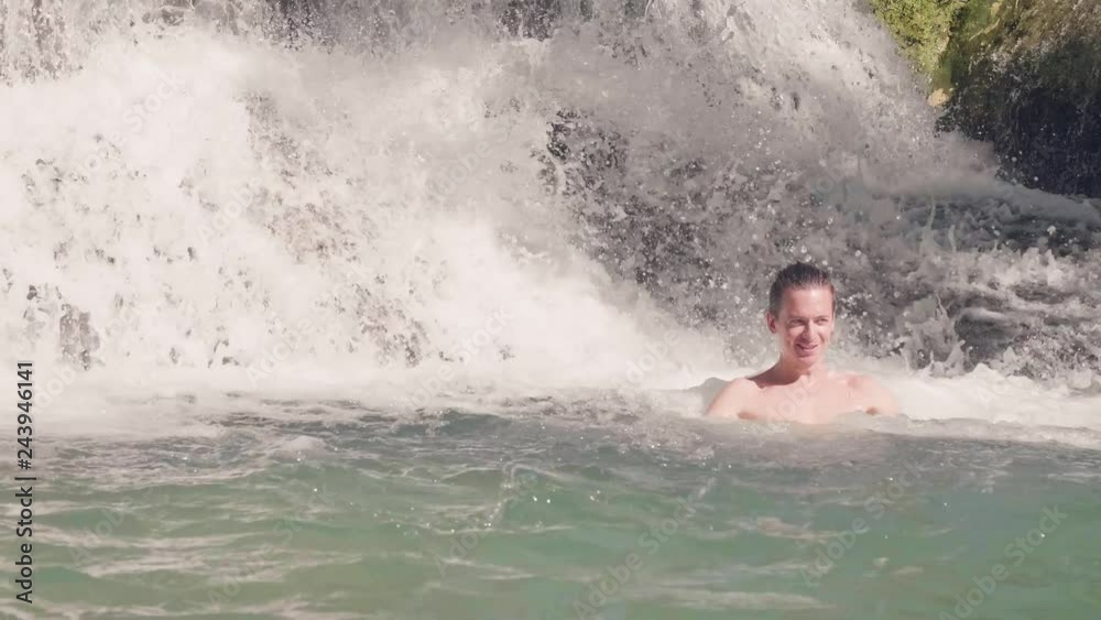 Young man swimming in water from splashing waterfall in tropical jungle. Smiling man bathing in flowing waterfall in mountain river. Splashing water stream from tropical river. Young man swimming in water from splashing waterfall in tropical jungle. Smiling man bathing in flowing waterfall in mountain river. Splashing water stream from tropical river.