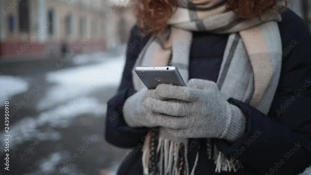 Close-up of young woman wearing warm scarf and gloves typing on touch screen of mobile phone outdoors in winter city.