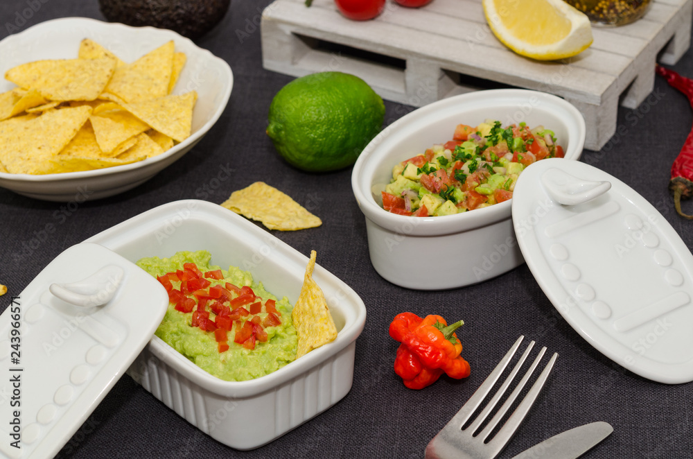 Guacamole and salsa dip next to tortilla chips. A delicious bowl of guacamole next to fresh ingredients on a table with tortilla chips and salsa. Guacamole and salsa dip next to tortilla chips. A delicious bowl of guacamole next to fresh ingredients on a table with tortilla chips and salsa.