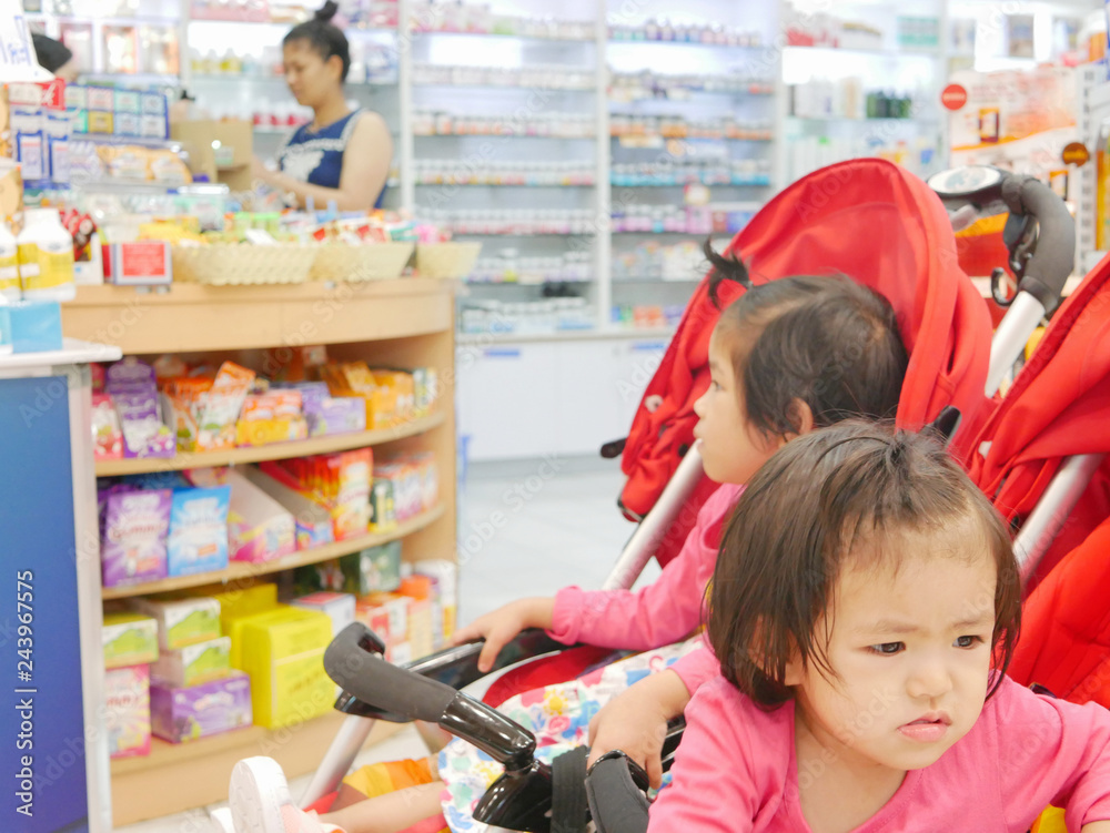 Little Asian baby girl (front), 22 months old, getting bored waiting for a long time in a baby stroller, with her older sister, for her mother buying drugs at a pharmacy