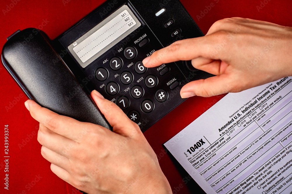 Human dials a phone number next to tax Form 1040X on a and red velvet background, close-up Human dials a phone number next to tax Form 1040X on a and red velvet background, close-up