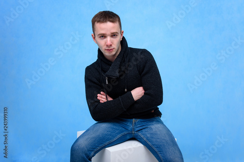 Concept studio portrait of a young man on a blue background sitting on a cube. © Вячеслав Чичаев
