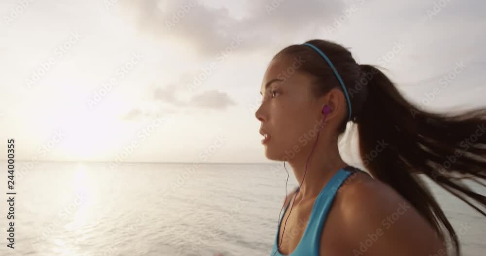 Determined female runner jogging at beach during sunset. Sporty young woman is listening music through earphones against sky wearing blue sports bra. SLOW MOTION, STEADICAM, RED EPIC.