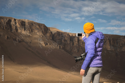 Portrait from the back of a traveling girl in a down jacket with a cap taking pictures of an epic landscape with rocks on her smartphone © yanik88